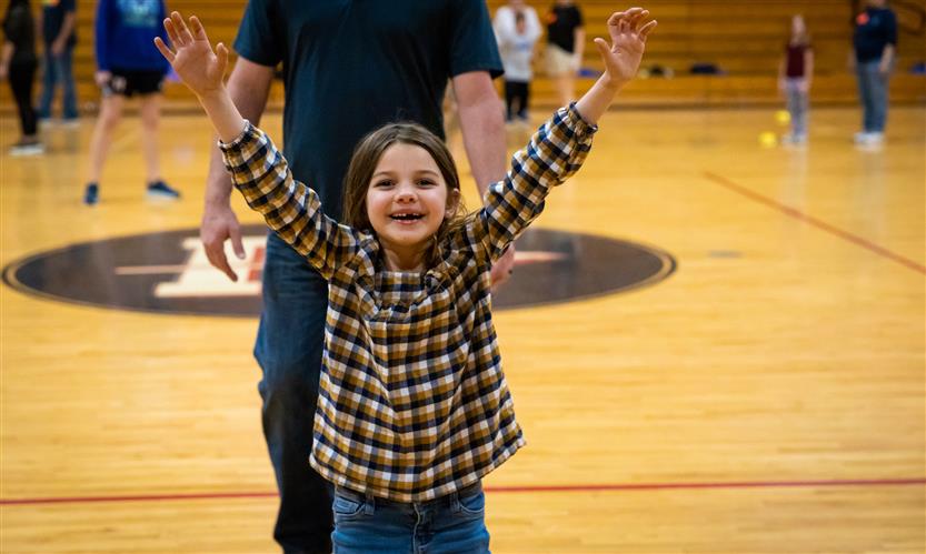 A girl with long, brown hair jumps for joy in a school gym